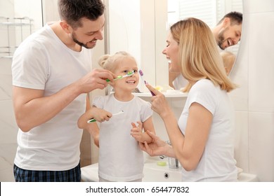 Young Couple And Their Beautiful Daughter Brushing Teeth Near Mirror In Bathroom