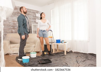 Young Couple Thinking At Living Room Renovation Sitting In The Middle Of The Room. Home During Renovation, Decoration And Painting. Interior Apartment Improvement Maintenance. Roller, Ladder For House