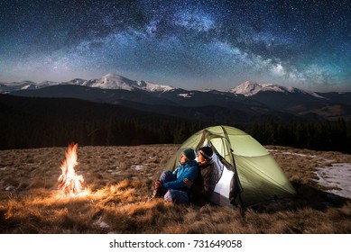 Young Couple Tourists Enjoying In The Camping At Night, Having A Rest Near Campfire And Green Tent Under Beautiful Night Sky Full Of Stars And Milky Way. On The Background Snow-covered Mountains