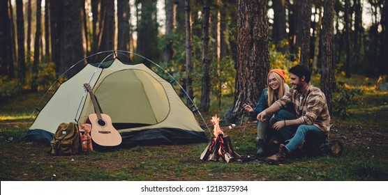 Young couple of tourists are exploring new places. Attractive woman and handsome man are spending time together on nature. Sitting near bonfire and touristic tent in forest and making marshmallow. - Powered by Shutterstock