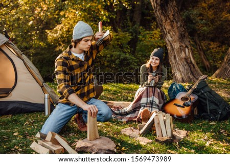 Young couple of tourists are exploring new places together. Handsome man chopping wood with axe. Attractive woman drinks tea and sitting on the log. Trevel concept. Boyfriend is tired from job.