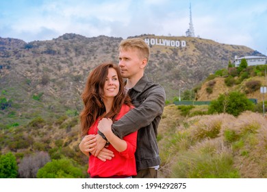 A Young Couple Of Tourists, A Man And A Woman, Embrace In Hollywood Against The Backdrop Of A Hill With The Famous Sign, Los Angeles