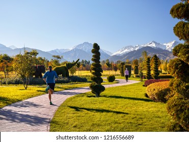 Young Couple Training In Topiary Park. Man Is Jogging And Woman Doing Nordic Walking With Poles. Healthy Life Concept. 