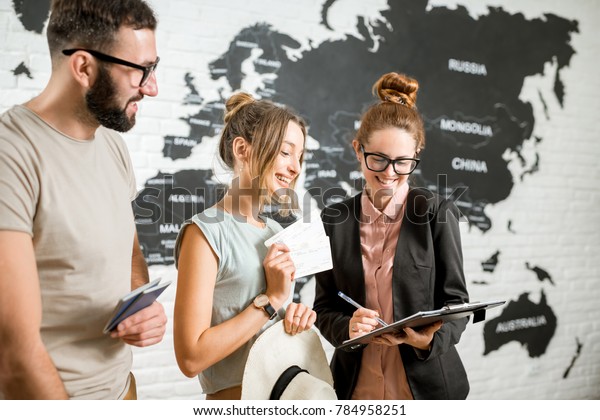 Young couple of\
travelers choosing a summer strip standing with travel agent on the\
world map background