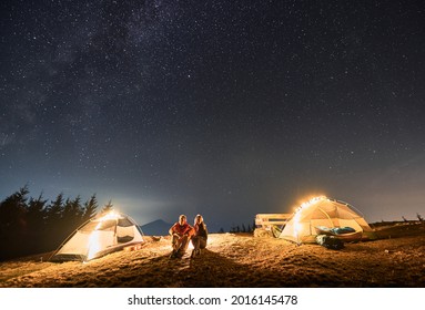 Young Couple Travelers Sitting On Grassy Hill Near Tourist Tents And Looking At Majestic Night Sky With Stars. Concept Of Hiking, Night Camping And Relationships.