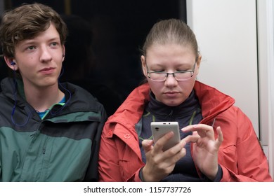 Young Couple Is Travelling In London Underground. He Listens To Music, She Surfs On Her Smartphone.