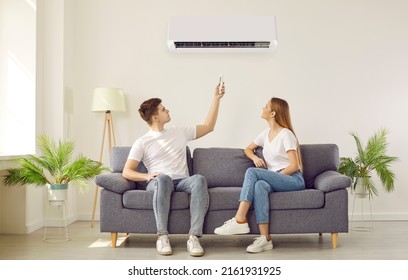 Young couple turn on air conditioner at home. Man and woman sitting on sofa in cozy white living room interior use remote to change AC temperature settings. Domestic air conditioning in house concept - Shutterstock ID 2161931925