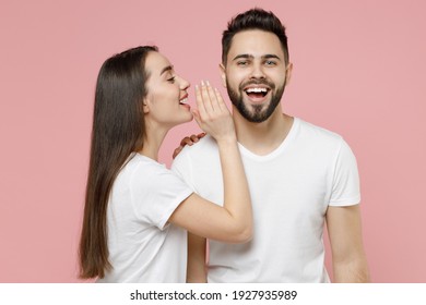 Young couple two friends bearded man brunette woman in white basic blank print design t-shirts whispering gossip and tells secret behind her hand, sharing news isolated on pastel pink color background
