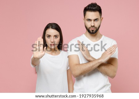 Young couple two friends bearded man woman in white basic blank print design t-shirts say no holding palm folded crossed hands in stop gesture isolated on pastel pink color background studio portrait