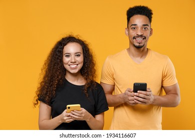 Young couple two friends together family african man woman 20s in black t-shirt holding using mobile cell phone chatting in social network dating isolated on yellow color background studio portrait