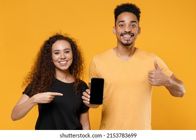 Young Couple Two Friends Together Family African Man Woman In Black T-shirt Hold Mobile Phone, Blank Screen Workspace Area Show Thumb Up Like Gesture Isolated On Yellow Background Studio Portrait