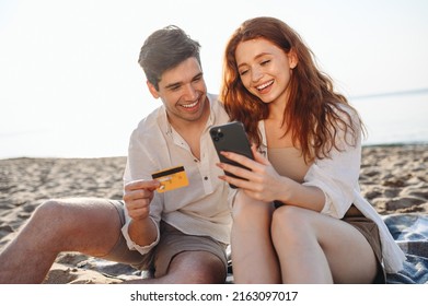 Young Couple Two Man Woman 20s In White Clothes Sit On Sand Use Mobile Phone Credit Card Shopping Online Order Delivery Book Tour Rest Together At Sunrise Over Sea Beach Outdoor Seaside In Summer Day