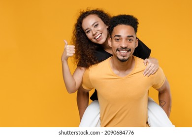 Young Couple Two Together Family Smiling Happy African Man Woman 20s In Black T-shirt Giving Piggyback Ride To Joyful Sit On Back Show Thumb Up Gesture Isolated On Yellow Background Studio Portrait