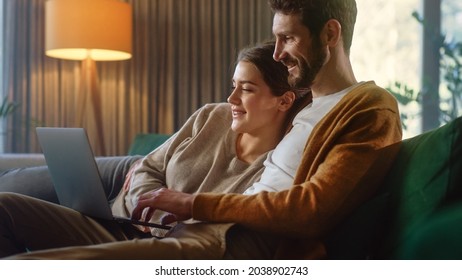 Young Couple Use Laptop Computer, While Sitting On A Couch In The Cozy Stylish Apartment. Boyfriend And Girlfriend Shopping On Internet, Using Social Media, Watching Funny Videos And Streaming Service