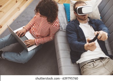 Young Couple Using Electronic Devices. Man Playing Breathtaking Computer Game With VR Headset. Woman Typing On Laptop. Modern Family Life