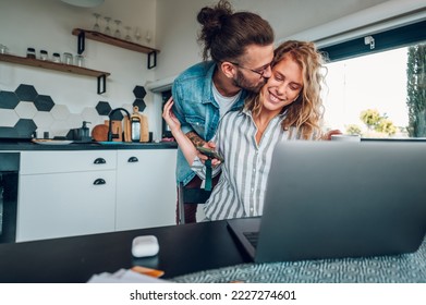 Young couple using laptop in the kitchen at home while smiling woman working online on a laptop and her husband is kissing her. Family morning lifestyle with technology concept. Copy space. - Powered by Shutterstock