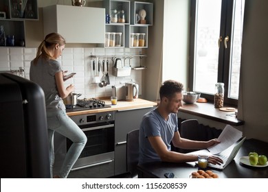 Young Couple Using Laptop And Smartphone In The Kitchen, Smiling Man Working Online At Home While His Wife Cooking With Phone Apps, Family Morning Lifestyle With Gadgets Or Devices Addiction Concept