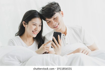 Young Couple Using Phone In Bed With Cheerful Expressions