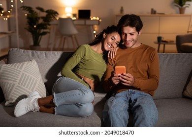 Young Couple Using Phone Browsing Internet Relaxing Sitting On Sofa At Home At Night. Husband And Wife Texting Via Cellphone Or Checking New Mobile Application On Smartphone Indoor. Modern Gadgets