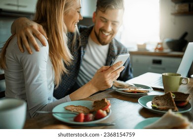 Young couple using a smartphone while having breakfast together in the morning in the kitchen at home - Powered by Shutterstock