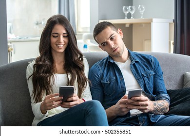 Young Couple Using Smartphones At Home. Jealous Boyfriend Spying His Girlfriend's Phone.