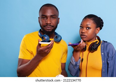 Young Couple Using Speaker On Mobile Phone To Record Message, Wearing Headset Over Blue Background. Boyfriend And Girlfriend Holding Mobile Phone To Use Microphone, Making Speech Online.