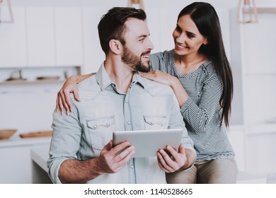 Young Couple Using Tablet PC in White Kitchen at Home. Breakfast at Morning. Happy Wife and Husband. Romantic Relationship Concept. Smiling Young People at Home. Using Digital Device.