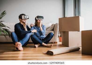 A Young Couple With VR Goggles Sitting On A Floor, Moving In A New Home.