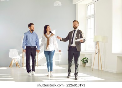 Young Couple Walking About A Big House Or Apartment Together With A Real Estate Agent Who Is Giving Them A Tour, Showing Around And Telling About The Advantages Of Living Here. Buying House Concept