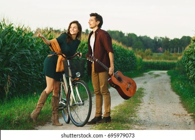 Young couple is walking by country road. Girl riding bicycle and guy is holding guitar.

