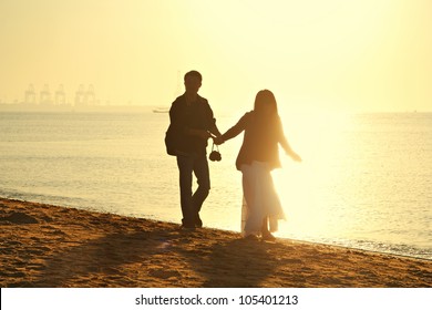 Young Couple Walking On The Beach At Dawn. Shooting Backlit. There Is A Port Qinhuangdao In The Distance.