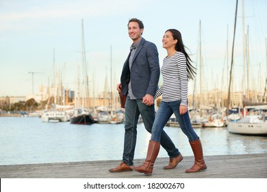 Young Couple Walking Outdoors In Old Harbor, Port Vell In Barcelona Catalonia, Spain. Romantic Happy Woman And Man Holding Hands Enjoying Life And Romance Outside. Multiracial Caucasian Asian Couple.