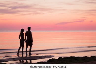 Young Couple Walking At The Seaside In Summer Evening. Man's And Woman's Silhouettes On The Beach. Sea Water Reflecting A Pink Heavenly Colors And Creating A Romantic Atmosphere.