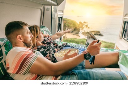 Young couple watching a movie on the tablet eating popcorn lying on the bed of their camper van with the doors open - Powered by Shutterstock