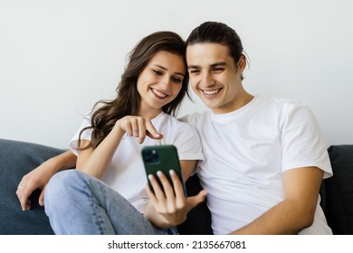 Young Couple Watching Online Content In A Smart Phone Sitting On A Sofa At Home