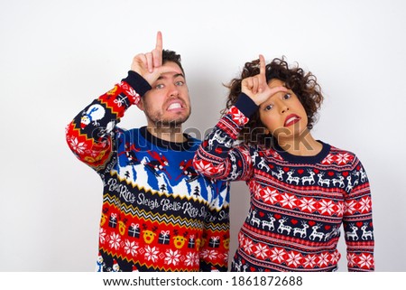 Young couple wearing Christmas sweater standing against white background making fun of people with fingers on forehead doing loser gesture mocking and insulting.
