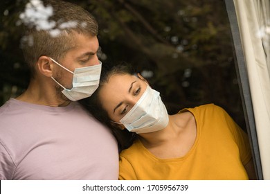 Young Couple Wearing Face Mask In Front Of The Window And Cuddling Each Other At Home. Coronavirus And Quarantine Concept