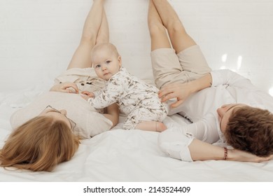 Young couple in white clothes lying with legs raised straight up high on white bed with awesome chubby blue-eyed baby infant toddler sitting between parents, posing. Bedtime, relaxation, family, love. - Shutterstock ID 2143524409
