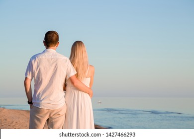 Young couple in white clothes. Man hugging woman at beach in summer evening. Orange sunset light. Back view. Empty place for inspirational, sentimental text, positive, lovely quote or cute sayings. 