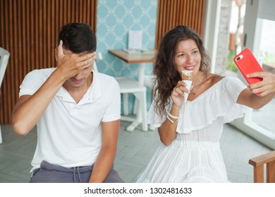 Young Couple In White Is Sitting In Cafe. Man Unhappy That Woman Takes Selfie Photo On Phone While Eating Ice-cream. Man Closes His Eyes With Hand.