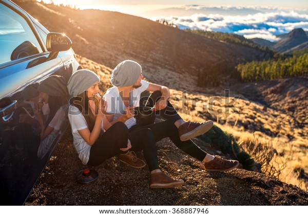 Young couple in white t-shirts and hats having\
picnic with croissant and cooking pan near the car on the mountain\
roadside above the clouds