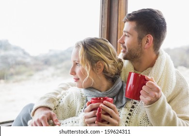 Young Couple In Winter Clothing With Coffee Cups Looking Out Through Cabin Window