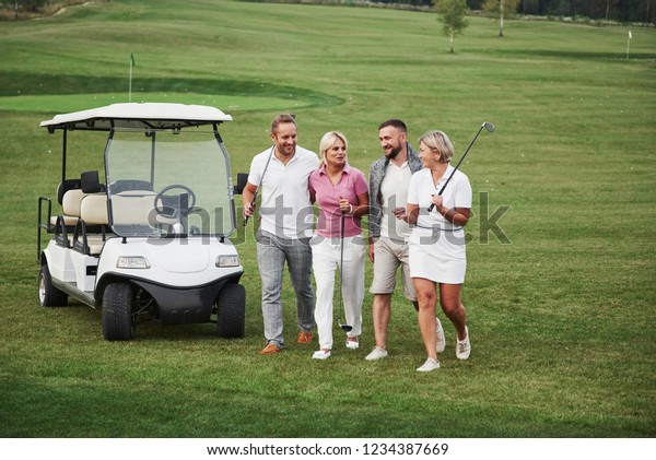 Young couples getting ready to\
play. A group of smiling friends came to the hole on a golf\
cart.