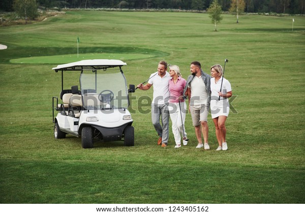 Young couples getting ready to\
play. A group of smiling friends came to the hole on a golf\
cart.