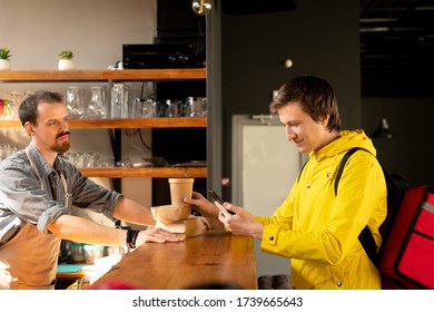 Young courier with backpack looking at client address in smartphone and checking order while standing by counter in front of waiter