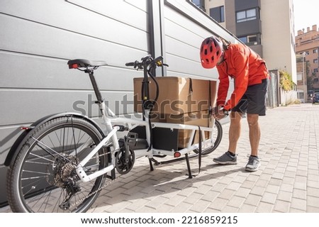 young courier with red clothing and helmet riding cargo bike arriving at the shipping destination to deliver a package to a city address, unloading the cargo