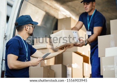 Young courier using touchpad while loading packages with his coworker in a delivery van. 