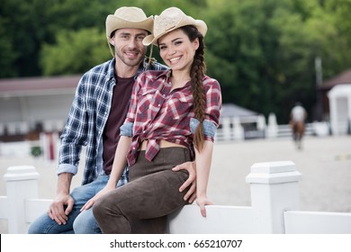 Young Cowboy Style Couple Embracing And Looking At Camera While Sitting On Fence