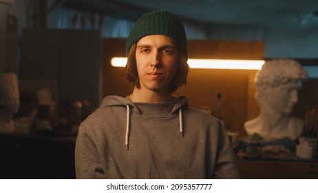 Young Craftsman In Hoodie And Hat Looking At Camera While Standing In Dimly Illuminated Sculpting Studio