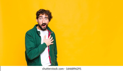 Young Crazy Bearded Man Feeling Shocked And Surprised, Smiling, Taking Hand To Heart, Happy To Be The One Or Showing Gratitude Against Flat Wall